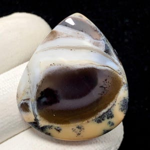 Puede incluir: Una gema pulida en forma de lágrima. La piedra presenta una mezcla de colores, incluyendo blanco, marrón y negro, sobre una base beige crema. El patrón evoca un paisaje con bandas superpuestas e inclusiones oscuras en forma de árbol. Mide aproximadamente 4 cm.