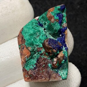 May include: A close-up of a natural mineral specimen. The stone displays a mix of colours, including deep blue, vibrant green, and earthy brown. The mineral's surface has a textured appearance, with visible crystalline structures.
