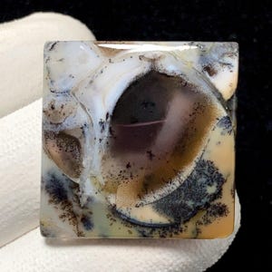 Puede incluir: Gema cuadrada con un patrón único. La piedra presenta una mezcla de colores, incluyendo blanco, marrón, negro y beige. El diseño se asemeja a un paisaje con formaciones arbóreas y patrones arremolinados. La piedra está pulida y tiene un acabado brillante.