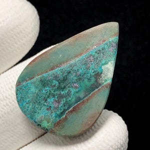 May include: A teardrop-shaped gemstone with a mix of teal, brown, and purple hues. The stone has a polished surface with a layered appearance, showcasing bands of color. The gemstone is held by a white glove.
