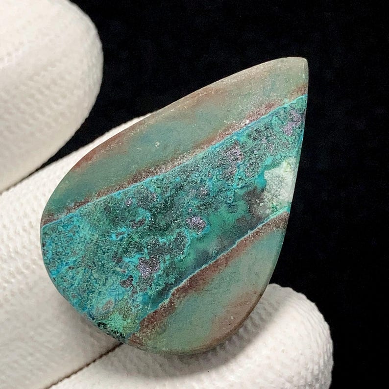May include: A teardrop-shaped gemstone with a mix of teal, brown, and purple hues. The stone has a polished surface with a layered appearance, showcasing bands of color. The gemstone is held by a white glove.
