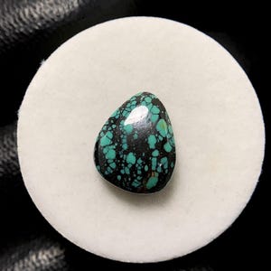 May include: A polished, teardrop-shaped gemstone with a striking pattern of turquoise-blue spots on a dark, almost black background. The stone is set on a white, circular surface, creating a high-contrast display. The gemstone's surface reflects light, highlighting its smooth texture.