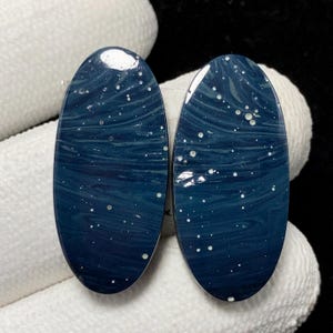 May include: Two oval-shaped cabochons with a deep blue, swirling pattern and scattered white speckles, resembling a starry night sky. The polished stones are displayed against a white background, highlighting their glossy surface and unique design.