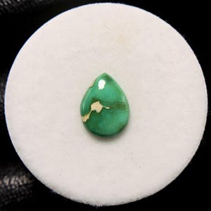 Puede incluir: Una gema pulida en forma de lágrima, de color verde vibrante con vetas blancas y marrones cremosas. La piedra se encuentra sobre un fondo circular blanco, ideal para la creación de joyas.