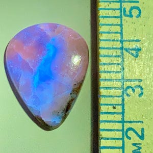 May include: A teardrop-shaped gemstone with a mix of pink, blue, and white hues. The stone has a polished surface and a dark brown edge. A ruler is visible in the background, with markings in inches.