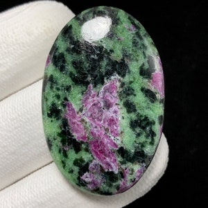 Op de afbeelding: Ovale edelsteen met een mix van groene, zwarte en roze tinten. Het gepolijste oppervlak reflecteert licht, waardoor de natuurlijke patronen en texturen worden benadrukt. De kleuren van de steen creëren een opvallend contrast.