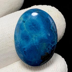 Peut inclure: Pierre précieuse ovale polie, d'un bleu profond, avec des inclusions bleu clair et marron foncé. La pierre est tenue sur un fond de tissu blanc, soulignant sa couleur et sa texture. La surface lisse du joyau reflète la lumière.