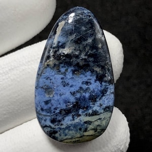 May include: A polished, teardrop-shaped gemstone with a blend of blue, black, and gray hues. The stone's surface has a marbled appearance, with swirling patterns and variations in color. The gemstone is held against a white background.