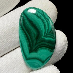 May include: Oval-shaped malachite gemstone with a polished surface. The stone features a vibrant green color with dark green, swirling patterns. The gemstone is held by a white gloved hand, with a black background.