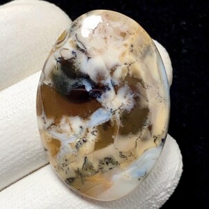 Puede incluir: Una gema ovalada con superficie pulida. La piedra presenta una mezcla de colores, incluyendo marrón, blanco, beige y toques de azul. El patrón se asemeja a un paisaje con vetas y remolinos, creando una apariencia natural única.