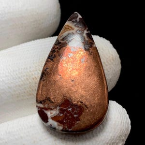 May include: A teardrop-shaped gemstone with a polished copper-colored surface. The stone features areas of white, brown, and black patterns near the top and bottom. The gemstone is likely used for jewelry or decorative purposes.