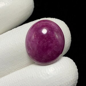 May include: A polished, oval-shaped gemstone in a deep magenta hue. The stone has a smooth, reflective surface with subtle internal patterns. The gem is held against a white background, highlighting its color and form. This gemstone could be used for jewelry.