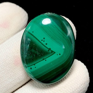 May include: Polished oval malachite gemstone. The stone displays a rich green hue with dark green and black banded patterns. The gemstone is held against a white cloth background.