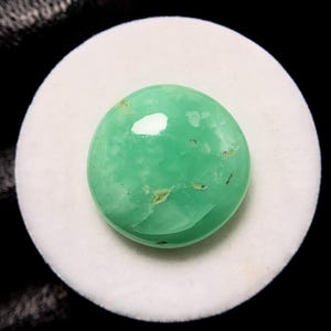 May include: A polished, oval-shaped, light green gemstone with minor inclusions. The gemstone is set on a white, circular surface, with a dark background. The stone has a glossy finish and a smooth texture.