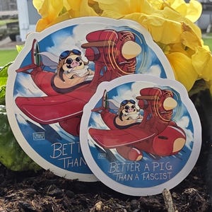 May include: Two round stickers featuring a cartoon pig piloting a red biplane against a blue sky with clouds. The stickers have the text "Better a Pig Than a Fascist". The pig wears goggles and a pilot's cap.