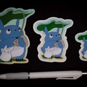 May include: Three stickers featuring a blue cartoon character holding a leaf umbrella, with a smaller white character. The stickers vary in size. A white and gray pen is in the foreground.