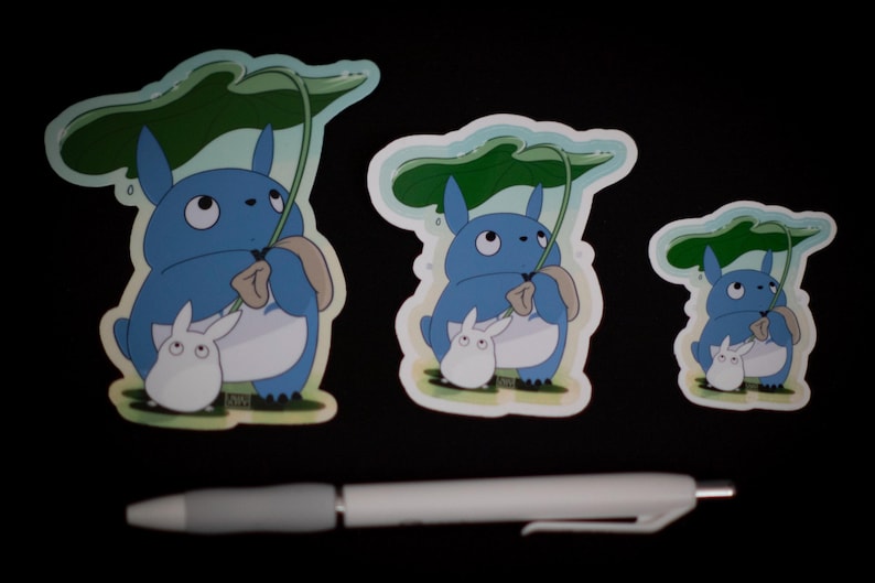 May include: Three stickers featuring a blue cartoon character holding a leaf umbrella, with a smaller white character. The stickers vary in size. A white and gray pen is in the foreground.