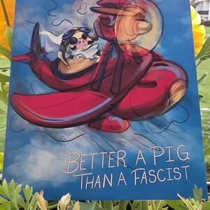 May include: A blue print featuring a cartoon pig wearing goggles and a scarf, flying a red airplane against a cloudy sky. The text "BETTER A PIG THAN A FASCIST" is written in white cursive.