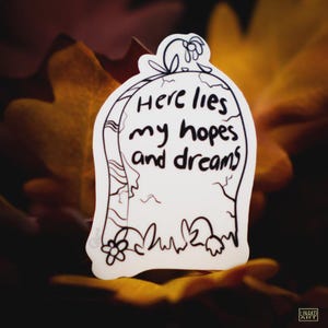 May include: A white sticker in the shape of a tombstone with the words "Here lies my hopes and dreams" in black lettering. The sticker has a black outline with floral and grass details. The background is blurred with fall leaves.