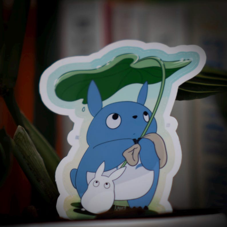 May include: A sticker featuring a blue cartoon character holding a large green leaf overhead. A smaller white character is next to the blue character. The sticker has a white border and a light blue background.