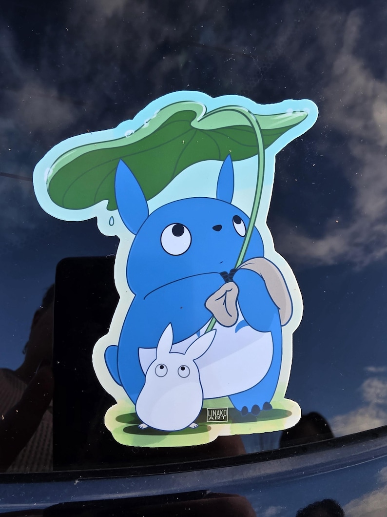 May include: A blue Totoro sticker with a white, smaller Totoro character. The larger Totoro holds a green leaf overhead. The sticker has a light blue outline and the artist's signature in the bottom right corner.