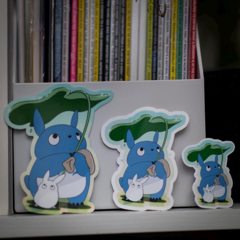 May include: Three stickers featuring a blue cartoon character holding a leaf umbrella, with a smaller white creature beside it. The stickers vary in size and are placed on a white surface. The background includes a shelf with books.