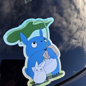 May include: A blue sticker featuring a cartoon character holding a green leaf umbrella, with a smaller white character beside it. The sticker has a light blue border and a grassy base.