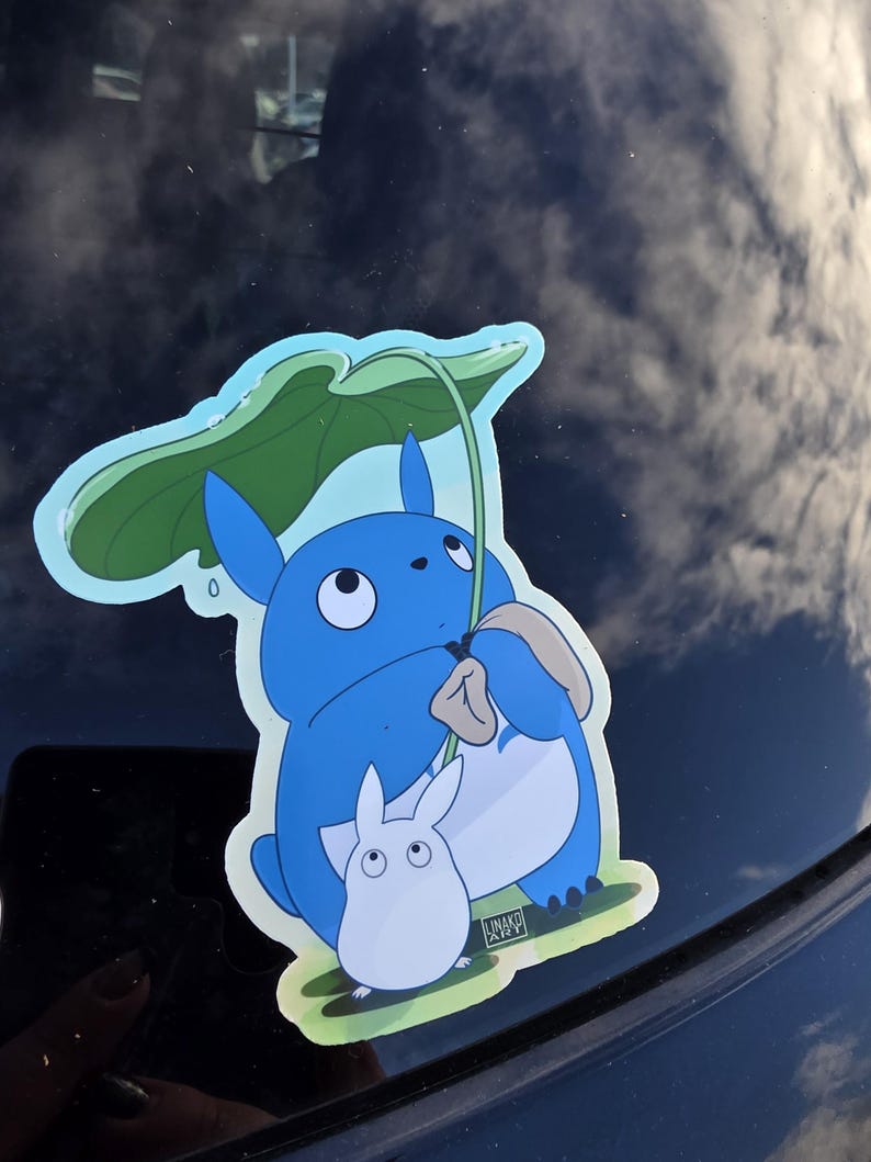 May include: A blue sticker featuring a cartoon character holding a green leaf umbrella, with a smaller white character beside it. The sticker has a light blue border and a grassy base.