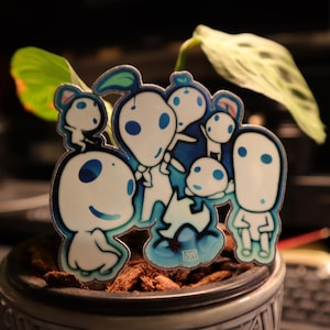 May include: A colorful sticker featuring a group of white, cartoon-like figures with blue accents. The figures have large, round eyes and are arranged in a cluster. The sticker is placed in a small pot with a plant.