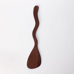 May include: A dark brown wooden spatula with a wavy handle and a wide, rounded head. The spatula is made of a single piece of wood and has a smooth finish. The design is modern and organic, suitable for kitchen use.