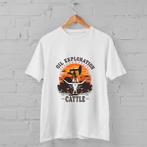May include: White t-shirt featuring a graphic with the text "OIL EXPLORATION CATTLE". The design includes an oil pump, a sunset, and a longhorn skull. The shirt is hanging on a wooden hanger.