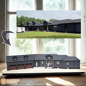 Könnte beinhalten: Ein Miniaturmodell eines dunkelgrauen Hauses mit einem passenden Bild des Hauses in Originalgröße im Hintergrund. Das Modell hat drei Garagentore mit braunen Zierleisten, Dachgauben und eine Familienfigur auf der Veranda. Das Logo "PAINTING STENCIL" ist sichtbar.