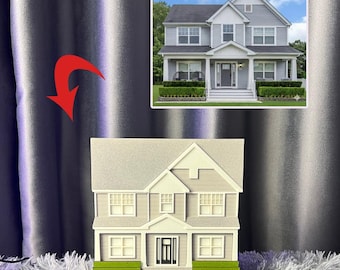 May include: A miniature house model with a gray and white color scheme, featuring a detailed facade with windows, a front door, and a small green hedge. The model is on a black base and has a matching image of the house in the background.