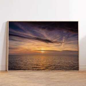 Può includere: Una fotografia incorniciata di un tramonto sull'oceano. Il cielo è un mix vibrante di viola, rosa e arancione, con nuvole vaporose. Il sole tramonta in lontananza, proiettando un bagliore dorato sull'acqua.