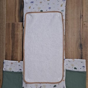 May include: A baby changing pad with a white towel center and a light brown border. The pad has a light blue background with a safari animal and palm tree pattern. Two green pockets are attached to the sides.