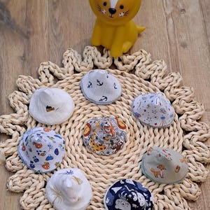 Peut inclure: Ensemble de bonnets pour bébés avec divers motifs, dont des ancres, des voitures et des animaux de la forêt, disposés sur un dessous de plat tressé. Une figurine de lion jaune est placée au-dessus. Les bonnets sont en tissu doux, pour bébés.