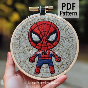 May include: Embroidered Spider-Man design in a wooden embroidery hoop. The superhero is depicted in red and blue, with white eyes and a black outline. The background is a light gray fabric with a spiderweb design. Text reads "PDF Pattern" and "Carmen Fortune".