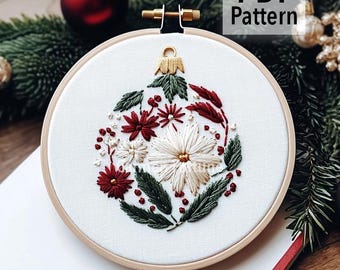 Könnte beinhalten: Ein Stickrahmen mit einem Christbaumkugel-Design. Die Kugel ist mit gestickten Blumen, Blättern und Beeren in Rot, Grün und Creme gefüllt. Der Text "PDF Pattern" ist sichtbar. Das Design befindet sich auf weißem Stoff in einem Holzrahmen.