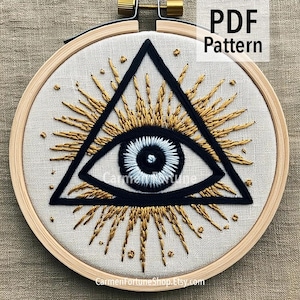 May include: Embroidered eye design in a wooden hoop. The eye is within a black triangle, surrounded by gold rays. The eye itself is white and blue. The text "PDF Pattern" is in the upper right corner.