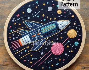 May include: Embroidered space-themed hoop art featuring a navy blue rocket ship with orange exhaust, surrounded by stars and planets. The text "PDF Pattern" is displayed at the top. The artist's name and shop URL are at the bottom.