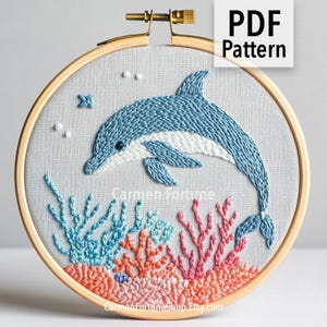 May include: Embroidered dolphin design in a wooden hoop. The dolphin is blue and white, swimming above coral and seaweed in shades of blue, orange, and pink. The text "PDF Pattern" is in the upper right corner.