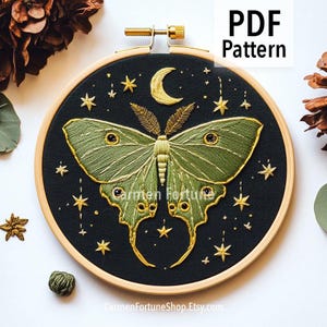 Könnte beinhalten: Gesticktes Motten-Design auf schwarzem Stoff, in einem Holzreifen. Die Motte ist grün mit goldenen Akzenten, umgeben von goldenen Sternen und einem Halbmond. Der Text "PDF Pattern" ist sichtbar.
