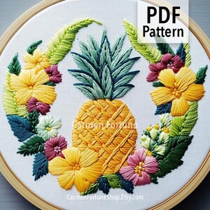 Op de afbeelding: Geborduurd ananasontwerp met gele, roze en groene bloemaccenten. De ananas is gecentreerd, omgeven door een krans van bloemen en bladeren. De tekst "PDF Pattern" staat in de rechterbovenhoek. Het ontwerp staat in een houten borduurring.