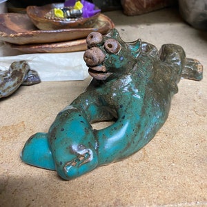 May include: A handmade ceramic sculpture of a mythical creature in shades of teal and brown. The creature has a large snout, multiple eyes, and a stylized body. The sculpture is resting on a wooden surface.