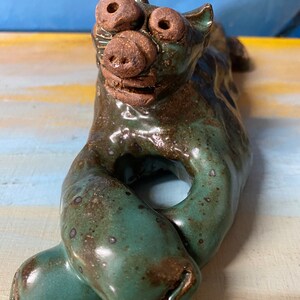 May include: A ceramic sculpture of a stylized animal with a pig-like snout and large, round eyes. The sculpture is primarily teal with brown accents and a glossy finish. The animal is in a reclining pose.
