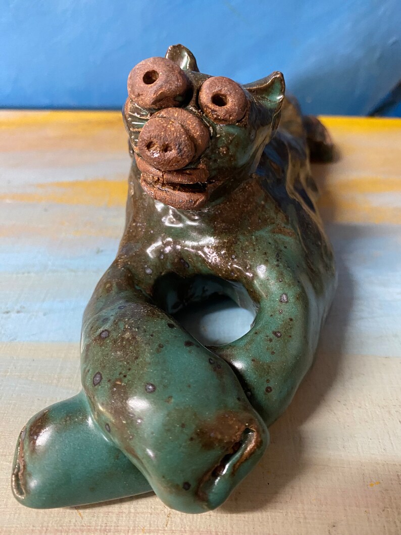 May include: A ceramic sculpture of a stylized animal with a pig-like snout and large, round eyes. The sculpture is primarily teal with brown accents and a glossy finish. The animal is in a reclining pose.