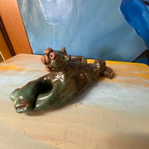May include: A handmade ceramic sculpture of a reclining animal, possibly a cat or dog, with a green and brown glaze. The sculpture is lying on its side, with its head and paws visible. The artwork is displayed on a painted surface.