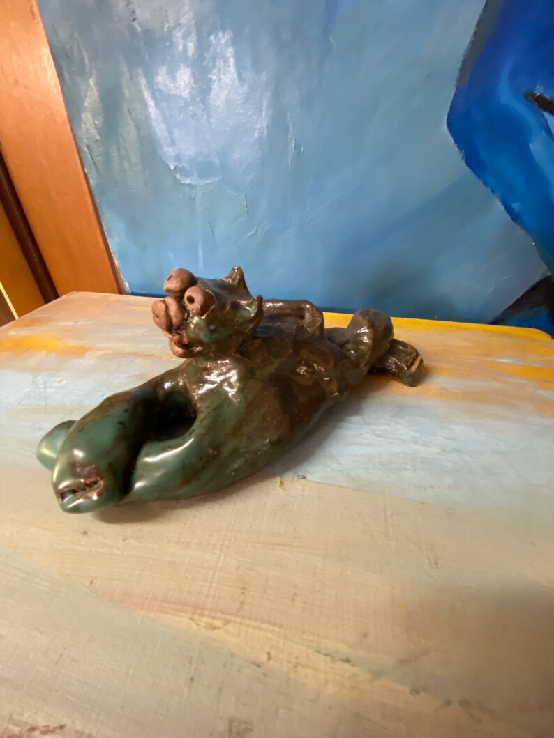 May include: A handmade ceramic sculpture of a reclining animal, possibly a cat or dog, with a green and brown glaze. The sculpture is lying on its side, with its head and paws visible. The artwork is displayed on a painted surface.