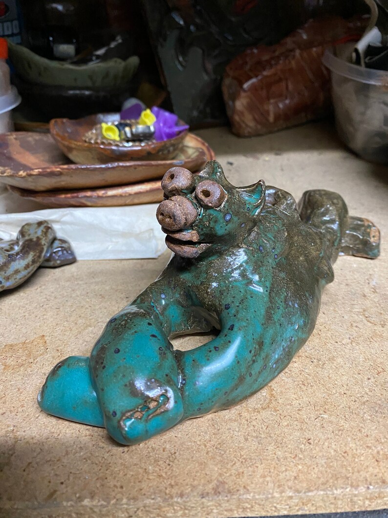 May include: A handmade ceramic sculpture of a mythical creature in shades of teal and brown. The creature has a large snout, multiple eyes, and a stylized body. The sculpture is resting on a wooden surface.