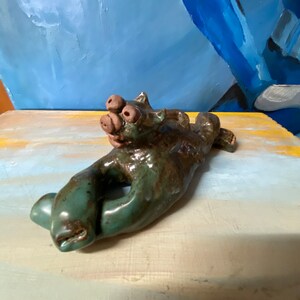May include: A handcrafted ceramic sculpture in the shape of a hand, with a creature-like head. The sculpture is glazed in shades of green and brown. The hand has a hole in the center. The head has three eyes and a snout.